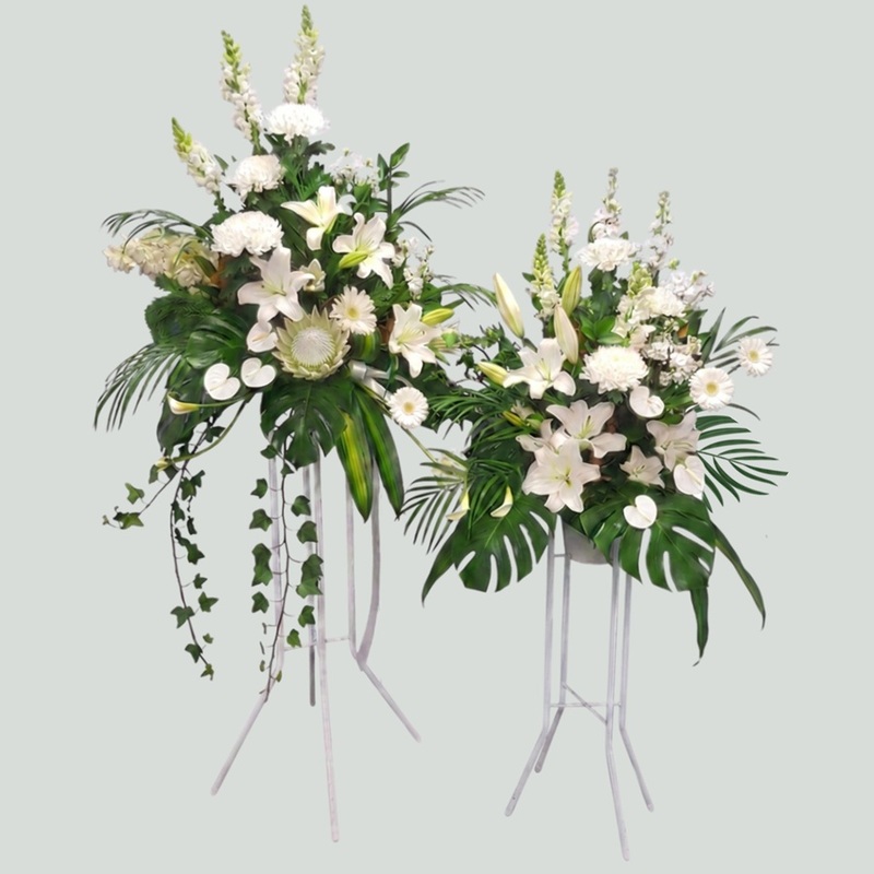 Coordinated pair of white standing funeral flower tributes with lilies, chrysanthemums, snapdragons and tropical foliage on tall and medium easel stands