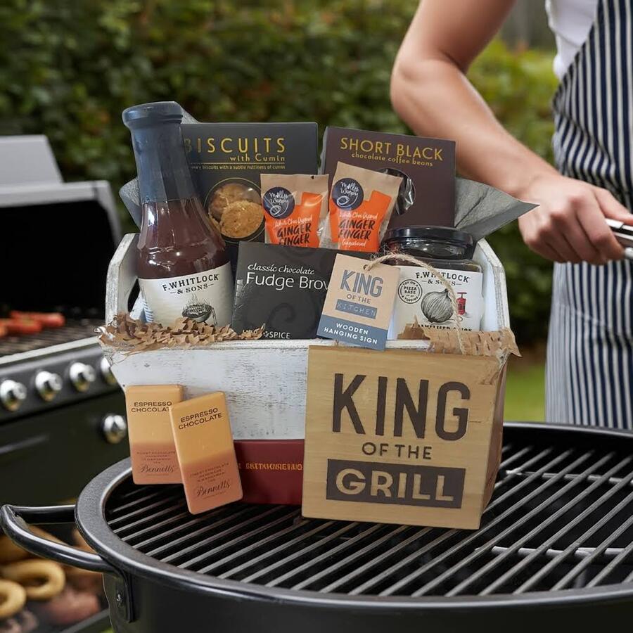 Gift Basket of BBQ themed items sitting on BBQ with Man in Apron beside Barbeque with sausages cooking.